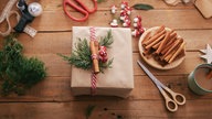 Ein Weihnachtsgeschenk in braunem Packpapier, verziert mit Tannengrün und Zimtstangen. Um das Paket herum auf dem braunen Holztisch liegen die Materialien, die zum Verpacken verwendet wurden. Am rechten Rand des Bildes steht außerdem eine Tasse Kaffee.