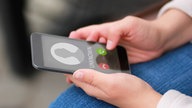 Ein Smartphone, auf dem gerade ein Anruf von unbekannter Nummer eingeht. Der Finger der Hand, die das Telefon hält, hovert unsicher über dem "annehmen" Button.