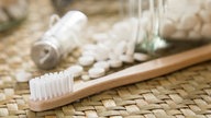 Eine Bambus-Zahnbürste, ein umgestoßenes Glas Zahnputz-Tabletten und eine Spule plastikfreie Zahnseide liegen auf einer geflochtenen Matte. 