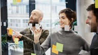 Lächelnde Arbeitskollegen in einem Büro schreiben auf eine Glastafel und kleben Haftnotizen daran. 