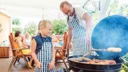 Ein Vater steht mit seiner kleinen Tochter an einem Grill und wendet ein Stück Fleisch.