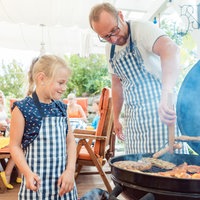 Ein Vater steht mit seiner kleinen Tochter an einem Grill und wendet ein Stück Fleisch.