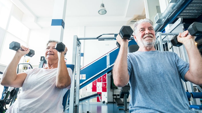 Eine Frau und ein Mann über 50 sitzen auf Fitnessgeräten und stämmen Gewichte