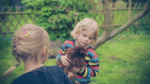 Zwei Kinder im Garten, eines von ihnen hält ein Huhn in den Händen