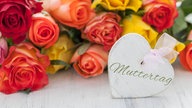 An einem Strauß mit roten, gelben und orangenen Rosen hängt ein Wweißes Herz, auf dem mit verschlungener Schrift "Muttertag" steht.