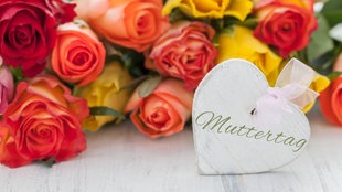 An einem Strauß mit roten, gelben und orangenen Rosen hängt ein Wweißes Herz, auf dem mit verschlungener Schrift "Muttertag" steht.