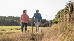 Ein Paar beim Nordic Walking an einem Feld mit einem Wald im Hintergrund