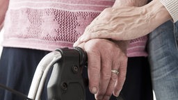 Nahaufnahme von Händen und dem Griff eines Rollators: Eine jüngere Frau unterstützt eine ältere Frau beim Gehen. 
