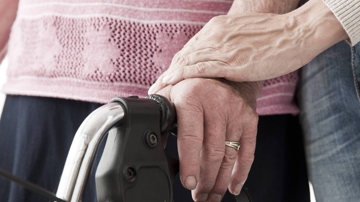 Nahaufnahme von Händen und dem Griff eines Rollators: Eine jüngere Frau unterstützt eine ältere Frau beim Gehen. 