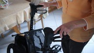 Eine ältere Frau läuft in ihrer Küche am Rollator. Ihre Hände an den Griffen sind im Fokus des Bildes, im Hintergrund sieht man ihren Küchentisch.