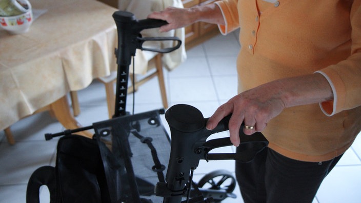 Eine ältere Frau läuft in ihrer Küche am Rollator. Ihre Hände an den Griffen sind im Fokus des Bildes, im Hintergrund sieht man ihren Küchentisch.