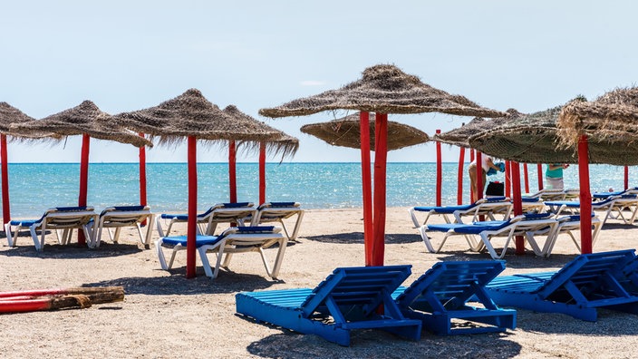 Liegestühle und Sonnenschirma an sonnigem Sandstrand mit Blick auf das Meer mit klarem, türkisem Wasser.