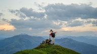 Ein Vater umarmt seine Tochter auf einer Bergspitze unter bewölktem Himmel und zeigt mit seiner Hand in die Ferne.