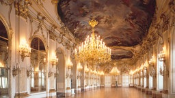Pompöse Galerie in einem Schloss mit bemalter Decke, goldenen Verzierungen und großen Kronleuchtern.