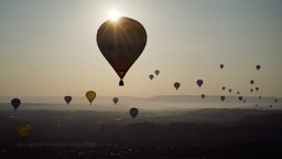 Viele Heißluftballons bei Sonnenaufgang über einer weiten Landschaft. Ein Ballon steht besonders im Vordergrund und wird von der Sonne angestrahlt, die anderen Ballons sind weiter weg am Horizont über Hügeln im Nebel zu sehen.