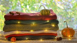 Ein gemütlich wirkendes, herbstliches Bild von einem Stapel Wolldecken, um den eine filigrane Lichterkette gelegt ist. Darauf und daneben befinden sich Zierkürbisse.