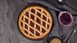 Von oben fortografiert: Eine Linzer Torte, ein traditionelles österreichisches Mürbeteiggebäck mit eingemachten Früchten und gehackten Nüssen mit Gittermuster. Neben der Torte stehen kleine Schalen mit den restlichen Früchten und  Nüssen.