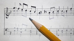 Ein Notenblatt mit zwei Notenzeilen. Die obere Zeile zeigt den Violinschlüssel, die untere den Bassschlüssel, anschließend ist jeweils eine Notenmelodie aufgezeichnet. Darüber wird ein Bleibstift gehalten, der die Noten zeichnet.