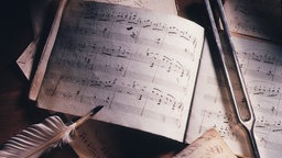 Ein aufgeschlagenes Notenheft, auf dem eine Stimmgabel und eine Schreibfeder liegen. 