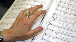 Nahaufnahme der Hand einer Frau, die in einer Partitur blättert. An einem Finger trägt sie einen auffälligen Ring. 
