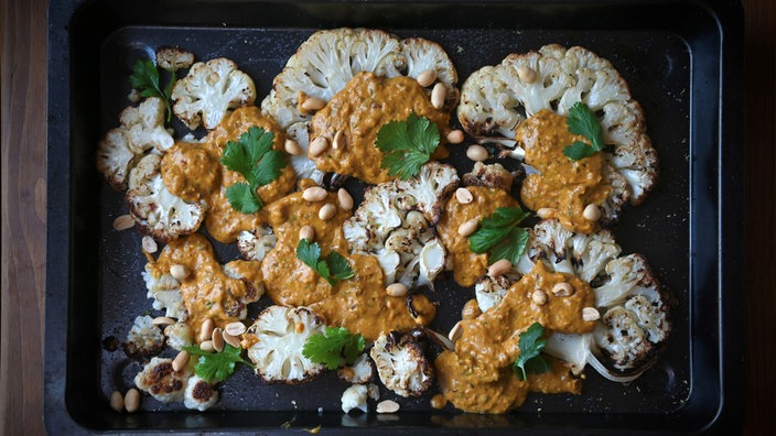 Von oben fotografiert: Eine Ofenform mit mehreren Scheiben Blumenkohl mit Kleksen Curry-Kokos-Sauce. Darüber gestreut sind Erdnüsse und Korianderblättchen. 