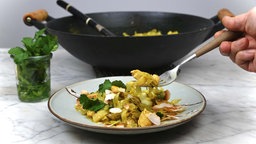 Spitzkohl und Hähnchen in Curry-Kokos-Sauce ist auf einem Teller angerichtet, im Hintergrund stehen ein Glas mit frischem Koriander und das restliche Curry in einer Wok-Pfanne