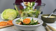 Eine Schale mit Salat, garniert mit einer Mandarinenscheibe. Im Hintergrund sieht man ein Brettchen mit Mandarinenschreiben, einen Kopf Eisbergsalat, eine große Schüssel mit dem restlichen Salat und eine Schale mit Käsesauce.