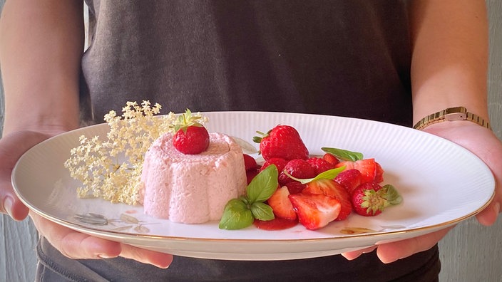 Eine Portion Erdbeermousse, daneben frische Erdbeeren garniert mit Basilikumblättern auf einem weißen Teller, der in die Kamera gehalten wird. Eine Holunderblüte zur Deko liegt ebenfalls daneben.