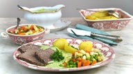 Im Vordergrund steht ein angrichteter Teller mit Rindfleisch, Kartoffeln und Leipziger Allerlei mit grüner Sauce. Im Hintergrund steht eine weitere Schale mit Leipziger Allerlei, eine Sauciere mit der Sauce, und eine Auflaufform mit Kartoffeln. 