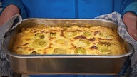 Eine silberne Ofenform mit Gratin, im Käse darauf sind Lauchstücke zu erkennen. man sieht rechts und links Ullas Hände, die die Ofenform mithilfe von Geschirrtüchern festhalten. 