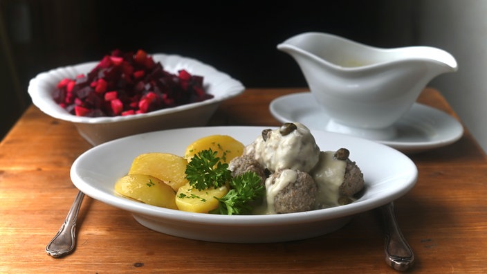 Ein angerichteter Teller mit Königsberger Klopse mit Soße übergossen und Kartoffeln, garniert mit Petersilie. Im Hintergrund steht eine Schale mit Rote Bete Salat und eine Sauciere.