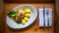 Auf einem Holztablett angerichtet: Ein Teller mit Kartoffeln und einer Kohlroulade, rechts daneben eine Serviette mit Gabel und Messer. Von oben fotografiert. 