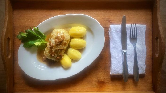 Auf einem Holztablett angerichtet: Ein Teller mit Kartoffeln und einer Kohlroulade, rechts daneben eine Serviette mit Gabel und Messer. Von oben fotografiert. 