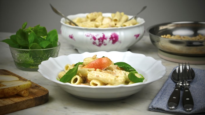 Auf einem gedeckten Tisch steht im Vordergrund ein tiefer Teller mit Pasta. Daneben liegen Gabel und Löffel, im Hintergrund sieht man eine große Schale mit noch mehr Pasta, eine Schale mit Basilikumblättern und eine Schale mit Mais. 