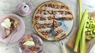 Von oben fotografiert: Auf einer Marmoroberfläche stehen ein angeschnittener Rhabarber-Pie, zwei Kuchenteller mit jeweils einem Stück des Pies, ein Brettchen mit weiteren Rhabarberstangen, und bereits geschnittenen Rhabarberstückchen.