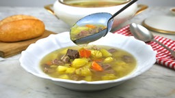 Ein Suppenteller mit Rindfleischsuppe, über den gerade eine gefüllte Suppenkelle mit noch mehr Suppe gehalten wird. Im Hintergrund sieht man ein Brötchen auf einem Brettchen, eine rot-weiß karierte Serivette mit einem Löffel und die Suppenterrine.