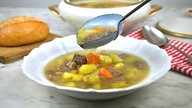 Ein Suppenteller mit Rindfleischsuppe, über den gerade eine gefüllte Suppenkelle mit noch mehr Suppe gehalten wird. Im Hintergrund sieht man ein Brötchen auf einem Brettchen, eine rot-weiß karierte Serivette mit einem Löffel und die Suppenterrine.