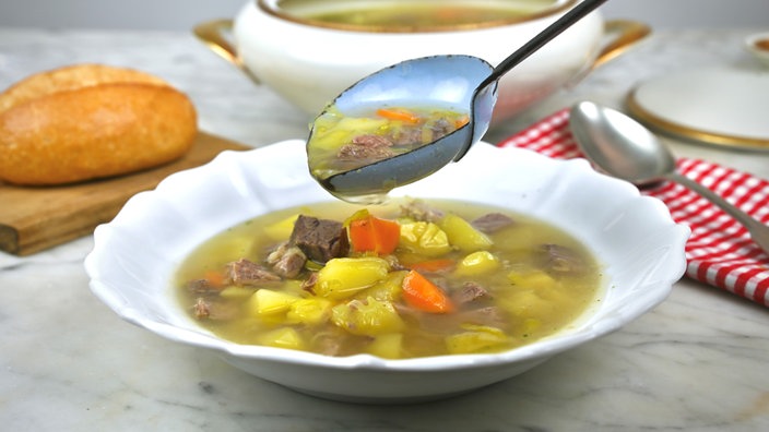 Ein Suppenteller mit Rindfleischsuppe, über den gerade eine gefüllte Suppenkelle mit noch mehr Suppe gehalten wird. Im Hintergrund sieht man ein Brötchen auf einem Brettchen, eine rot-weiß karierte Serivette mit einem Löffel und die Suppenterrine.