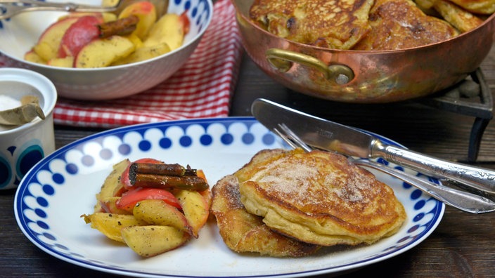 Zwei Pfannkuchen mit Bratkartoffeln und Paprikastreifen auf einem Teller, im Hintergrund eine Pfanne und Teller mit weiteren Pfannkuchen und Gemüse.