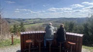 Zwei Menschen schauen von einem Aussichtspunkt auf Felder und Wälder