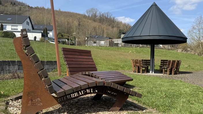 Zwei Liegen aus Holz auf einer Wiese. Daneben eine überdachte Sitzmöglichkeit