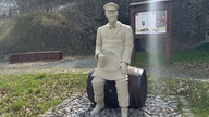 Eine weiße, meschengroße, männliche Figur, die mit einem Bierkrug auf einem Bierfass sitzt. Im Hintergrund Metallstelen mit Informationen