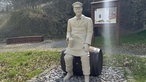 Eine weiße, meschengroße, männliche Figur, die mit einem Bierkrug auf einem Bierfass sitzt. Im Hintergrund Metallstelen mit Informationen