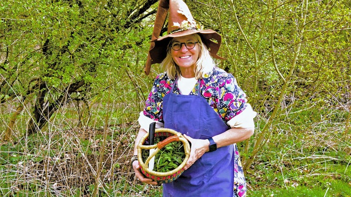 "Kraut Moni" steht mit einer Schürze, einem Hexenhut und einem Korb voll Kräuter am Wegrand und lächelt in die Kamera.