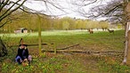 Monika Heimeier hockt mit Hut und Korb am Wegrand. Hinter ihr ist ein Wildgehege zu erkennen, in einiger Entfernung laufen zwei Rehe durch das Bild. 