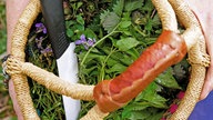 Nahhaufnahme eines Kräuterkörbchens von oben. In dem geflochtenen Korb befinden sich bereits abgeschnittene Kräuter und ein Messer. 