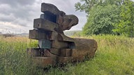 Ein Eichenstamm, der zu einer zweiteiligen Skulptur mit dem Titel "Reise und Heimkehr" aufbereitet wurde, liegt auf einer Wiese. 