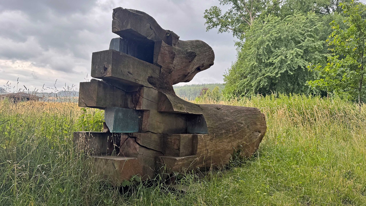 Ein Eichenstamm, der zu einer zweiteiligen Skulptur mit dem Titel "Reise und Heimkehr" aufbereitet wurde, liegt auf einer Wiese. 