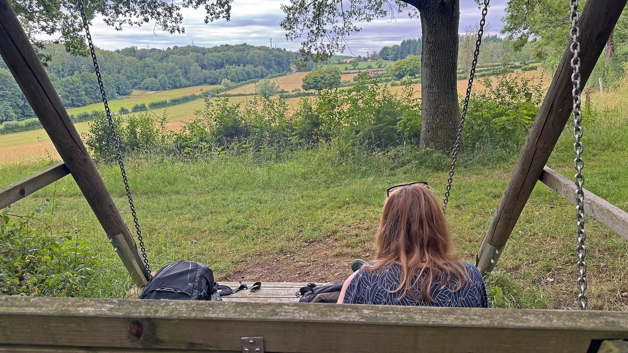 Eine Frau sitzt auf einer Hollywoodschaukel aus Holz und schaut auf ein weites Feld.
