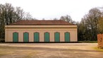 Langes, helles Gebäude mit rotem Ziegeldach und mehreren grünen Türen in einer offenen Parkanlage.